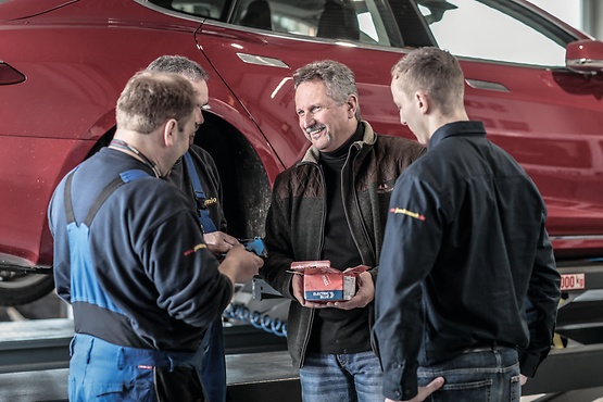 four mechanics talking about vehicle parts in front of car