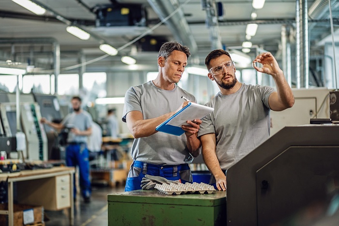TWO CNC machine operators examining finished products and writing notes in industrial facility.